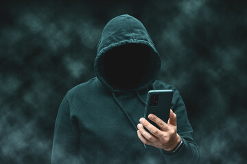 Mysterious faceless hooded anonymous hacker holding smartphone, silhouette of cybercriminal, terrorist