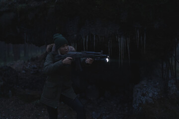 Fototapeta premium A civilian girl with a machine gun and a flashlight takes aim at night in the dark in the forest near the ruins, with weak lighting.