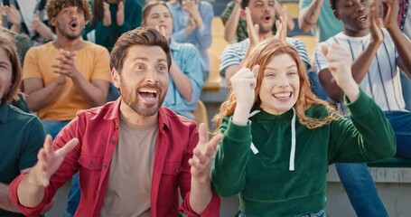 Happy young people watching football team playing at stadium. Positive fans loudly supporting players. Attractive guy hugging his girlfriend after team scoring goal. Happy crowd applauding winners.