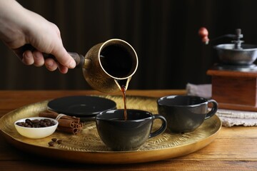 Turkish coffee. Woman pouring brewed beverage from cezve into cup at wooden table, closeup