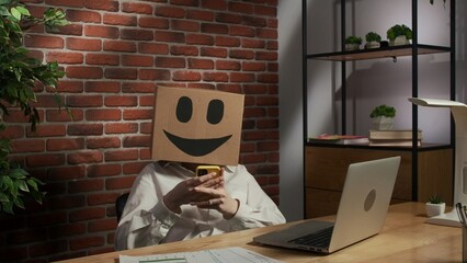 Portrait of female in cardboard box with emoji on head. Worker at the desk working on laptop,...