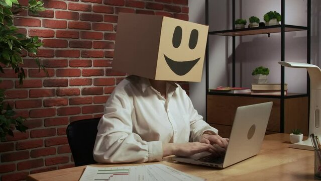 Portrait of female in cardboard box with emoji on head. Worker at the desk working on laptop, crossing hands looks at camera.