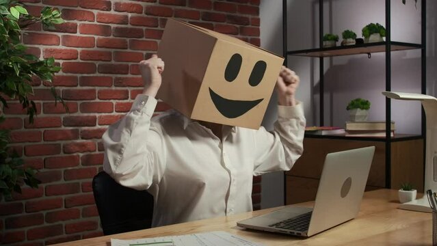 Portrait female in cardboard box with emoji on head. Worker at the desk working on laptop, winning gestures thumbs up at camera.