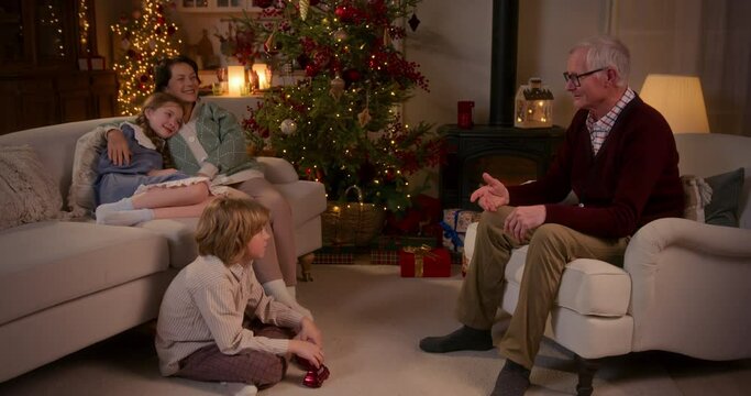 grandchildren sit with grandparents in living room near christmas tree at cozy home in the evening and listen a story told by grandfather. cuddling, smiling and feeling warmth