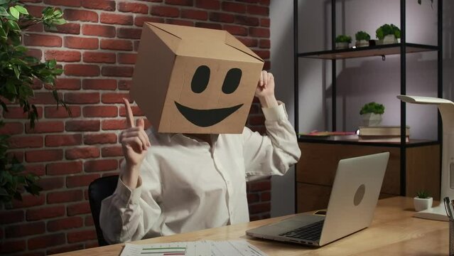 Portrait of female in cardboard box with emoji on head. Worker sitting at the desk looking at laptop, happy dancing positive mood.