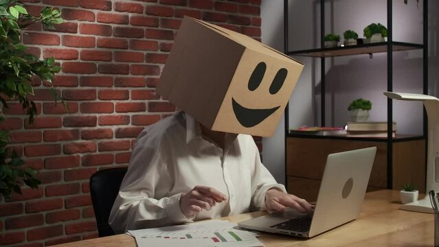 Portrait of female in cardboard box with emoji on head. Worker sitting at the desk working on laptop busy with paper documents.