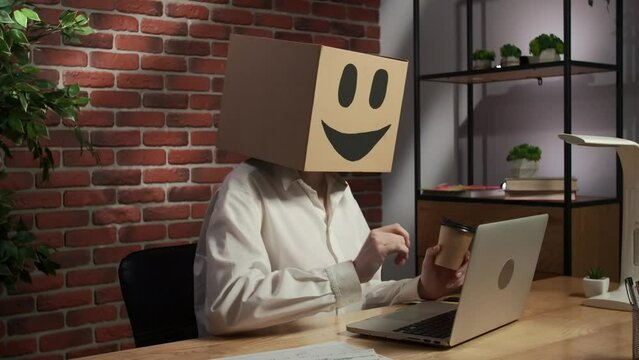 Portrait of female in cardboard box with emoji on head. Worker sitting at the desk working on laptop and drinking cup of coffee,