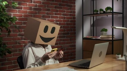 Portrait of female in cardboard box with emoji on head. Worker at the desk holding smartphone and credit card typing number.