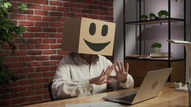 Portrait of female in cardboard box with emoji on head. Worker sitting at the desk talking by video call on laptop, happy face.