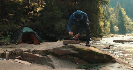 A lone tourist prepares food against the background of a camping site, a tent, a mountain river, a...