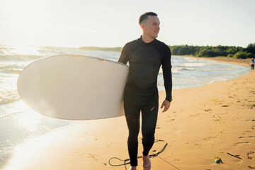 A male surfer walks along the beach holding a surfboard in a wetsuit.