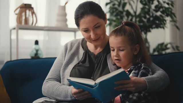 Mom and little daughter reading a book on couch at home, parenting, childhood