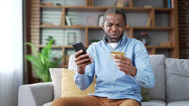 Frustrated mature african american man discovered fraud entering credit card number on smartphone sitting in room at home. Upset senior male became a victim of deception, money was stolen from account