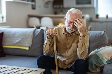 Depressed old man sitting at home while holding walking stick. Retired sad man holding wooden...