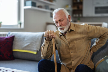 Senior man suffers from back pain. He sits on the sofa in the living room in one hand holding a walking stick while his other hand is on his painful back with a painful grimace on his face. - obrazy, fototapety, plakaty