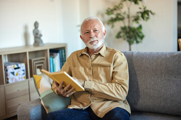 Fototapeta premium Bearded adult male holding interesting book while resting on the couch at home. Stylish mature man reading book after hard working day while sitting on cosy sofa. Domestic lifestyle concept