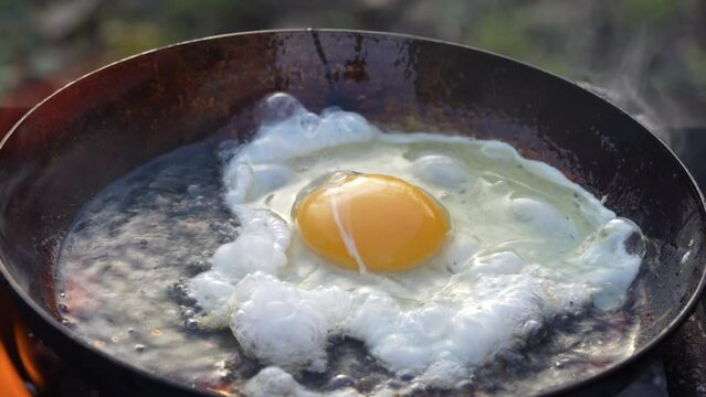 Fresh egg is fried in a frying pan outdoors with a sizzling oil and smoke on an open fire, close up