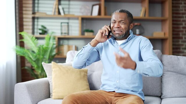 Angry excited mature african american male arguing on smartphone sitting on sofa in living room at home. Frustrated nervous senior black man complains on phone about problems that need to be solved