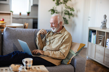 Elderly man with glasses with gray hair and beard sits at home on the sofa works or browses social...