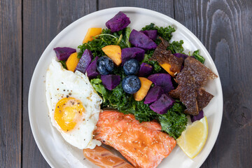 Healthy salad with fried egg, salmon, kale, sweet potato and blueberries on white plate.