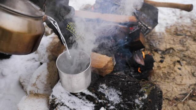 Unknown hiker in gloves pouring boiling water from aluminium kettle into metal cup outdoor. Traveler using kitchenware for heating water on flame to preparing warming drink while sitting by campfire.