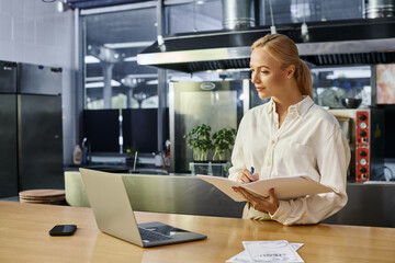 attentive blonde woman writing in notebook near laptop and smartphone on counter in modern cafe