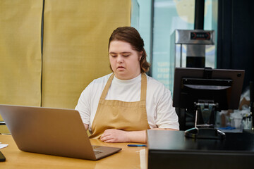 young female employee with mental disorder working on laptop near cash terminal on counter in cafe