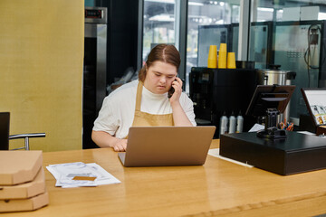 young woman with down syndrome talking on smartphone near laptop while working in modern cafe