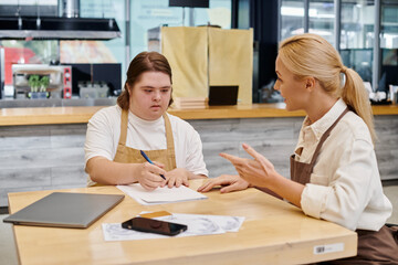 Fototapeta premium cafe administrator talking to female employee with down syndrome writing near laptop and smartphone