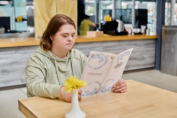 thoughtful female client with down syndrome looking at menu card while sitting at table in cafe