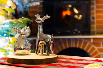 Two ceramic reindeer, silver and glossy, stand on a maple wood slice. In the background, a burning...
