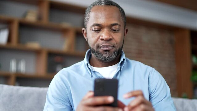 Mature african american bearded man using smartphone sitting on sofa in living room at home. Senior male browses social networks, does shopping in online store, chats, writes reads messages. Close up