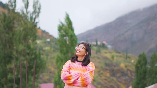 Smiling modern girl in warm sweater standing with crossed arms standing in mountains at Manali, Himachal Pradesh, India. Happy laughing cheerful female portrait having positive emotions. 