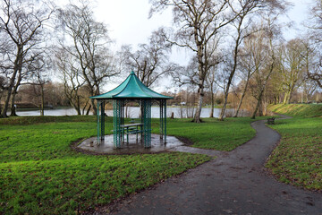 A green metal shelter covers a picnic seating area in a public park. Trees and grass are around it and a boating lake can be seen in the background. Shot in winter with a grey sky