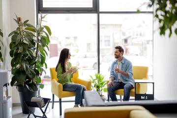 Cheerful adult man and woman talk while sitting in waiting room of a modern clinic. Young family waiting in comfortable lobby with greens, waiting for a doctor's appointment