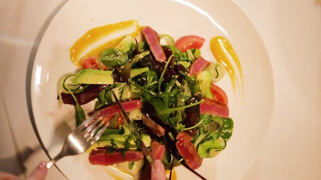 Top view of big plate with delicious tuna salad standing on white tablecloth in expensive restaurant. Close up of woman hands cutting piece of vegetable with knife and fork for enjoying tasty meal.