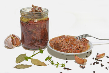 Canned stewed meat in a glass jar and on a plate, Nearby there is a fork for food and garlic, a bay leaf, black peas, all on a white background.