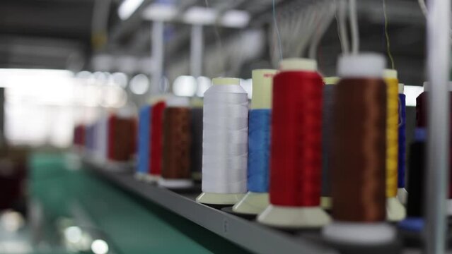 Multicolored coils of synthetic textile thread running at industrial weaving manufacturing machine. Selective focus on white sewing spool getting drawn at textile fabric. Concept of production.