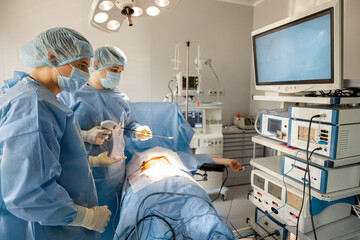 Two surgeons performs minimally invasive procedure with endoscopes, looking on monitor in operating room. Concept of endoscopic-computer assisted treatment - obrazy, fototapety, plakaty