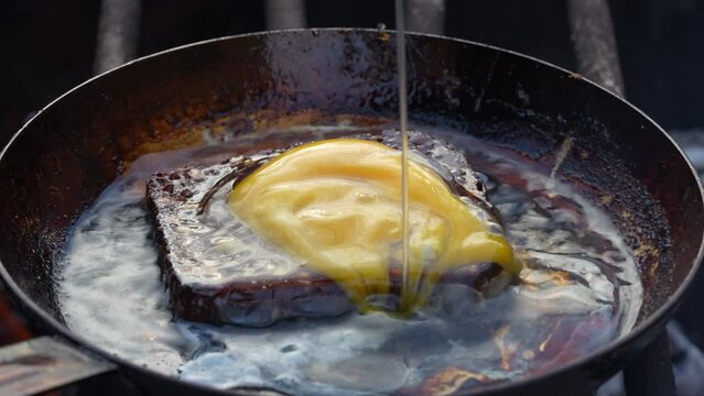 Piece of bread and fresh egg is fried in a frying pan outdoors with a sizzling oil and smoke on an open fire, close up