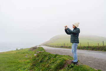 Latin woman with knit hat trying to take a photo on a foggy day on her car trip along the Irish coast