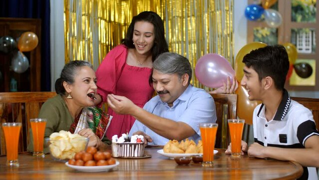 A cheerful middle-aged Indian man feeding a piece of cake to his wife - husband wife bonding  anniversary . An elderly couple and their grandchildren together - three generations  leisure time  bir...