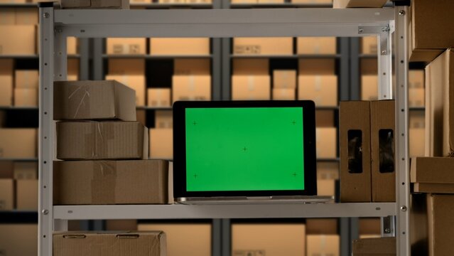 Laptop with chroma key green screen display standing on the storage rack shelf with boxes, workspace mockup advertising area.