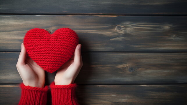 A red knitted heart in woman hands in red sweater on a wooden table background. Valentine's Day, love, romance concept. Top view, flat lay, copy space. Valentine's Day background.