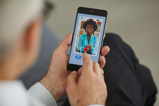Online dating concept. Closeup old man in search of love partner pressing red heart like button under young black woman's photo on dating website or mobile phone app. Hands holding cellphone close up