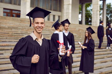 Portrait of happy male student with diploma on graduation day. Young man in black cap and gown...