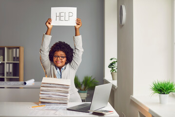 Asking help. Attractive african-american young woman holds piece of paper over her head with inscription help. Dark-skinned girl with pile of folders on her desk. Workplace in office with laptop. - obrazy, fototapety, plakaty