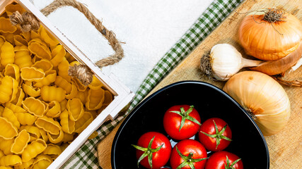 Top view of onions, garlic, pasta in a wooden crate and fresh ripe cherry tomatoes on a rustic white wooden table. Ingredients and food concept