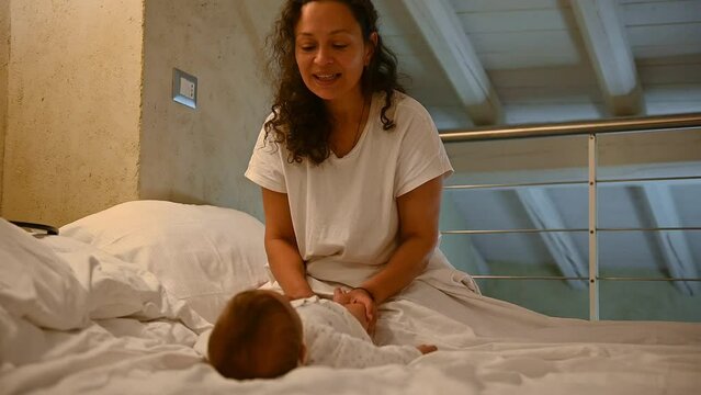 Slow motion. Latin American loving young mother sitting on the bed and performing gentle preventive massage and gymnastics to her baby boy, to prevent and reduce the risk of orthopedic diseases
