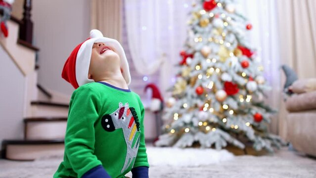 Joyful baby boy wearing red Santa cap pulling it on eyes. Happy toddler having fun at Christmas. Blurred backdrop.